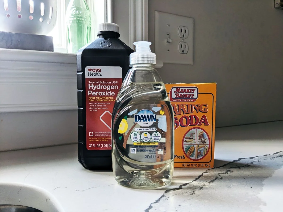 A bucket on a patio with hydrogen peroxide, baking soda, and dish soap set beside rubber gloves, ready for a skunk odor neutralizing wash
