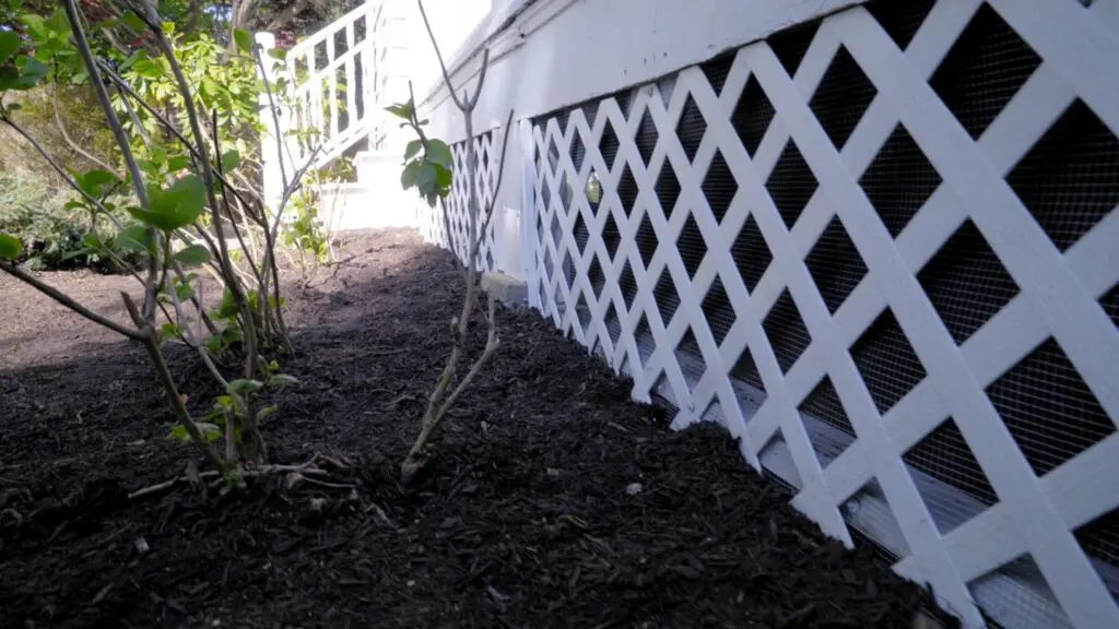 A person installing galvanized hardware cloth along the base of a wooden deck with a shallow trench dug for burial, realistic home improvement photo