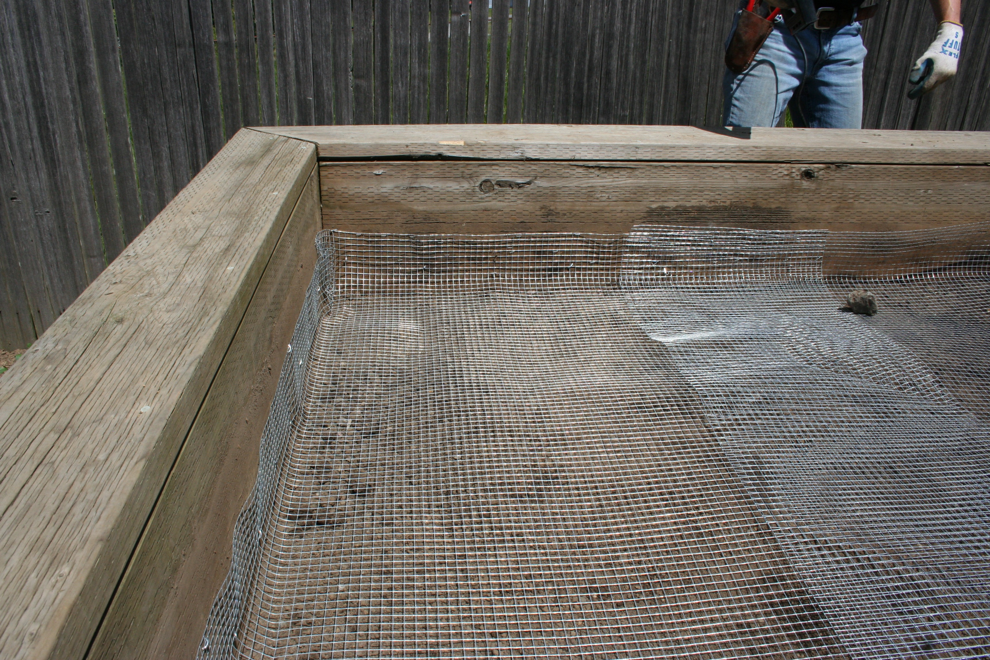 A raised garden bed under construction with galvanized hardware cloth stretched across the bottom frame before soil is added, photographed outdoors