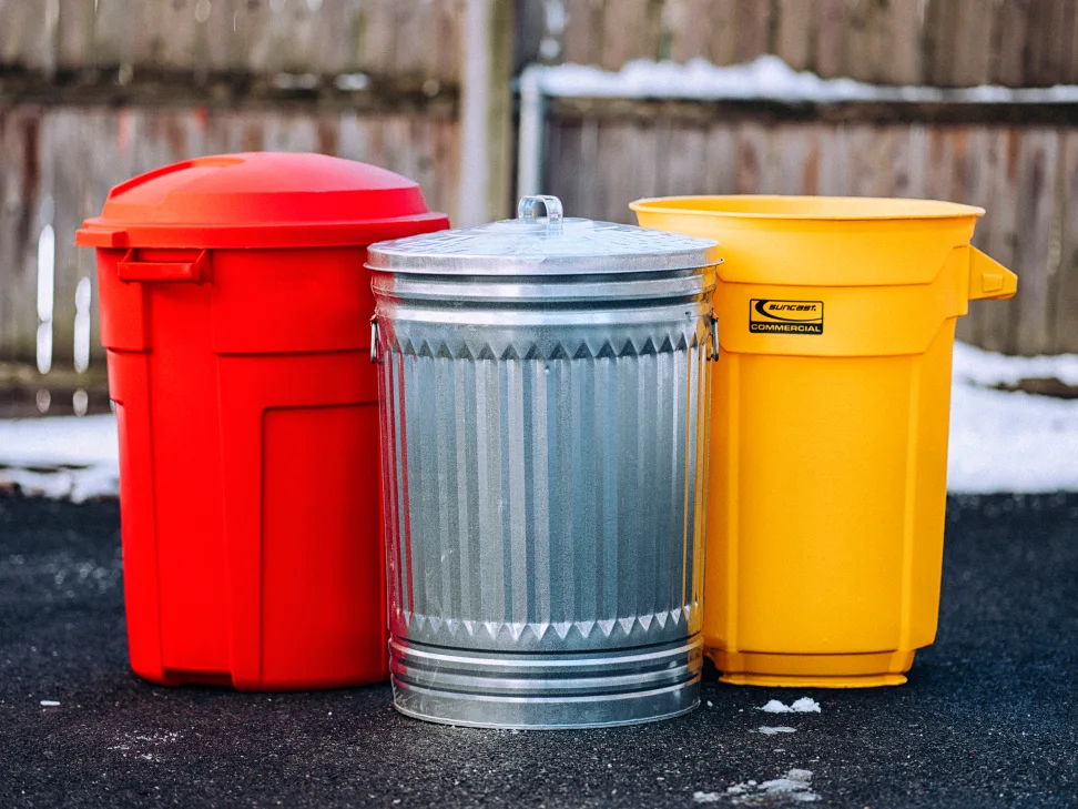 A real photo of two sealed metal trash cans with tight-fitting lids sitting on a clean concrete pad beside a backyard fence in daylight