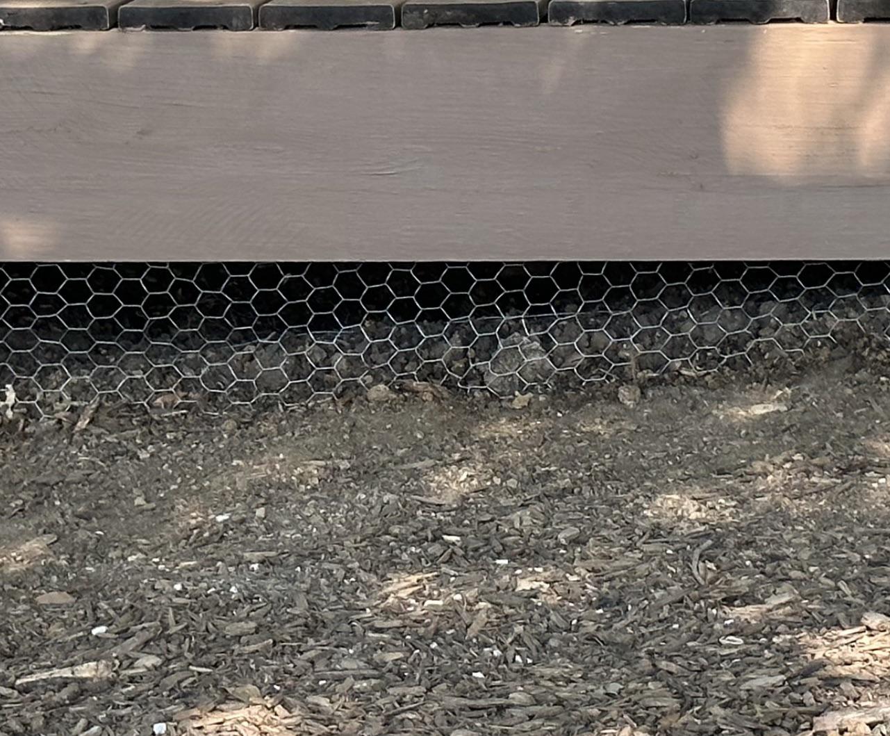 A real photo of welded wire mesh installed along the base of a wooden backyard deck, secured to prevent animals from digging underneath, with soil and mulch in the foreground, photorealistic