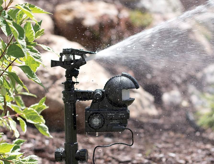 A real photograph of a motion-activated sprinkler installed in a garden bed near a fence line at dusk, with water spraying across a small area