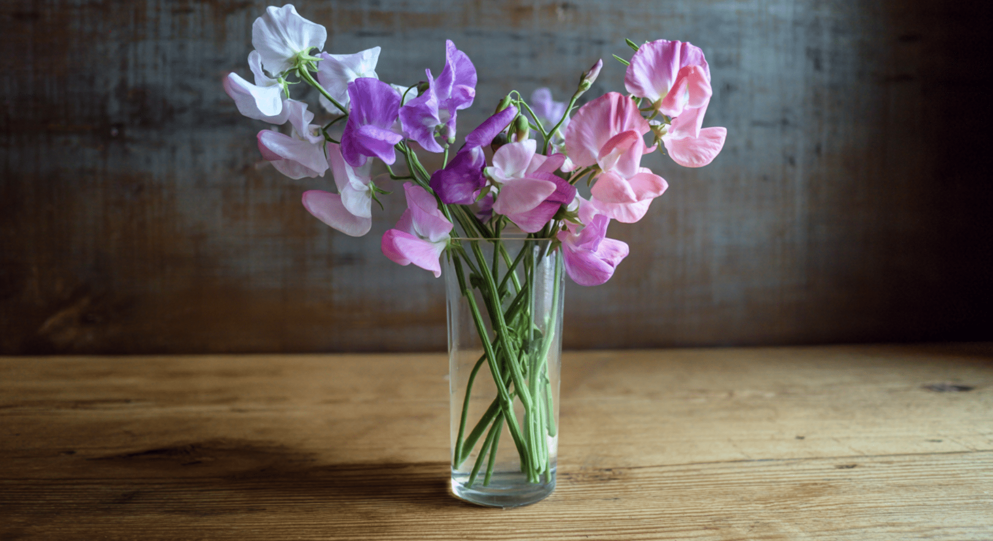 A real photograph of freshly cut sweet pea blooms in soft pastel shades arranged loosely in a clear glass vase on a wooden kitchen table