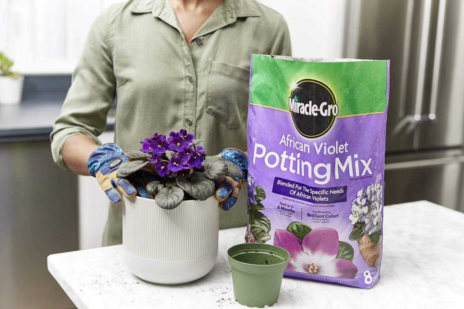 A small plastic nursery pot with drainage, fresh potting mix in a bowl, clean scissors, and a chopstick laid out on a table for repotting an African violet