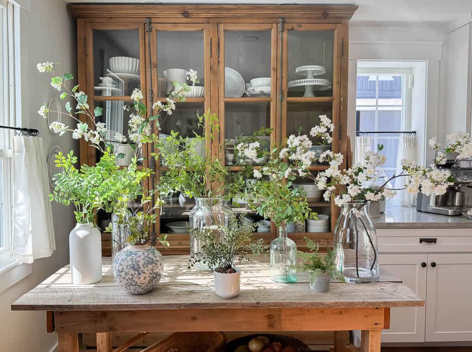 Flowering cherry branches with pale pink blossoms arranged in a clear vase on a wooden dining table with soft window light, realistic home photography style